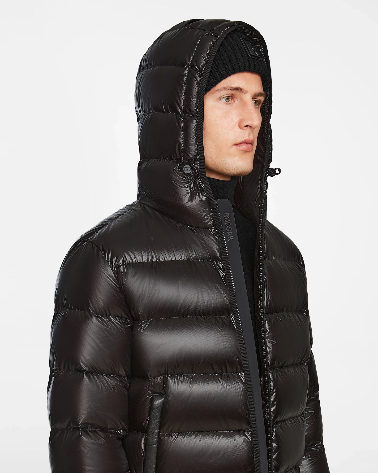 A person wearing the Rudsak DEMIAN S - M123508 Black mens down puffer jacket with the hood up and a black knit beanie, looking to the side against a plain white background.