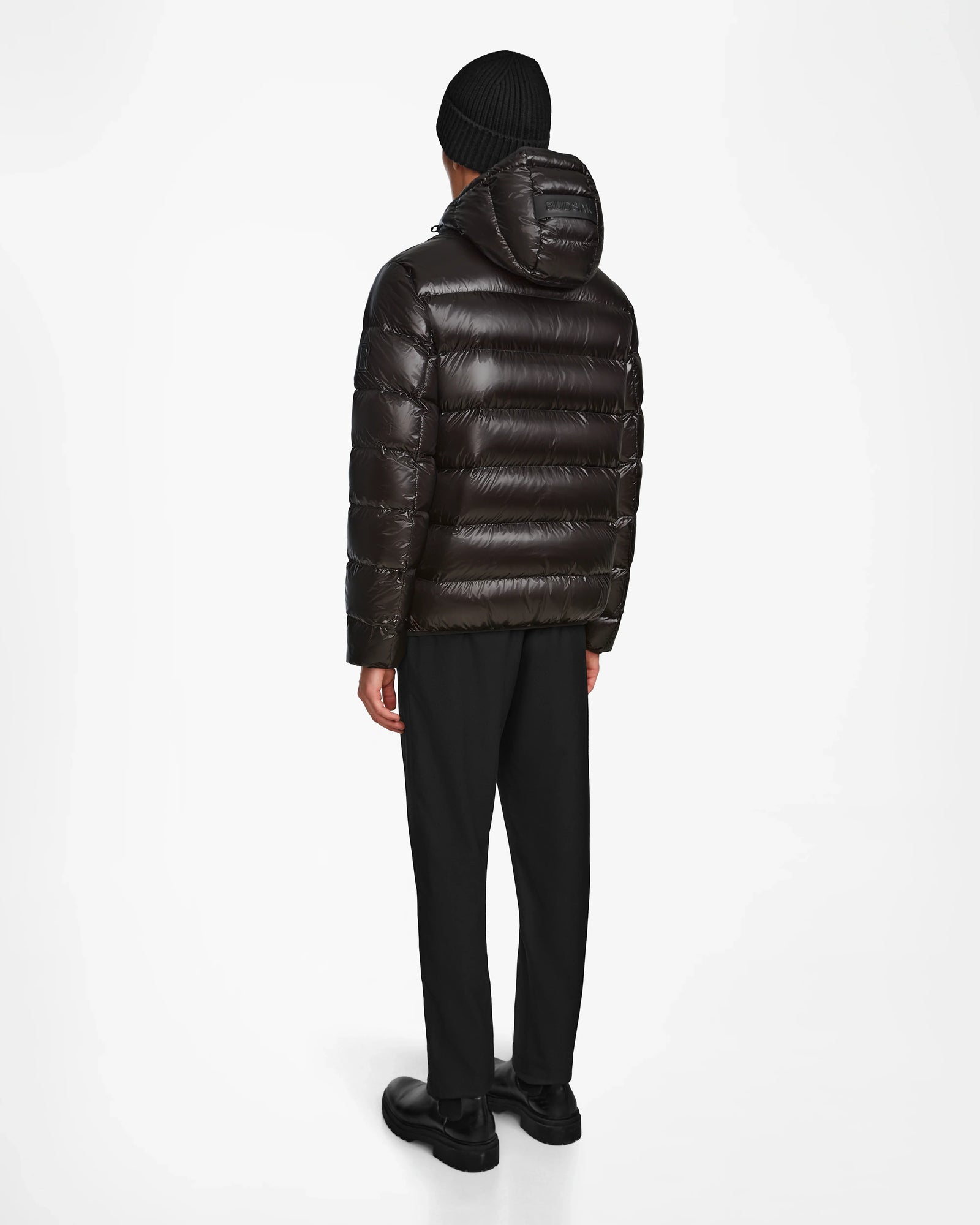 A person stands facing away in a Rudsak DEMIAN S - M123508 Black mens shiny down puffer jacket with hood, water- and wind-resistant, paired with black pants, shoes, and a knit beanie against a plain white background.
