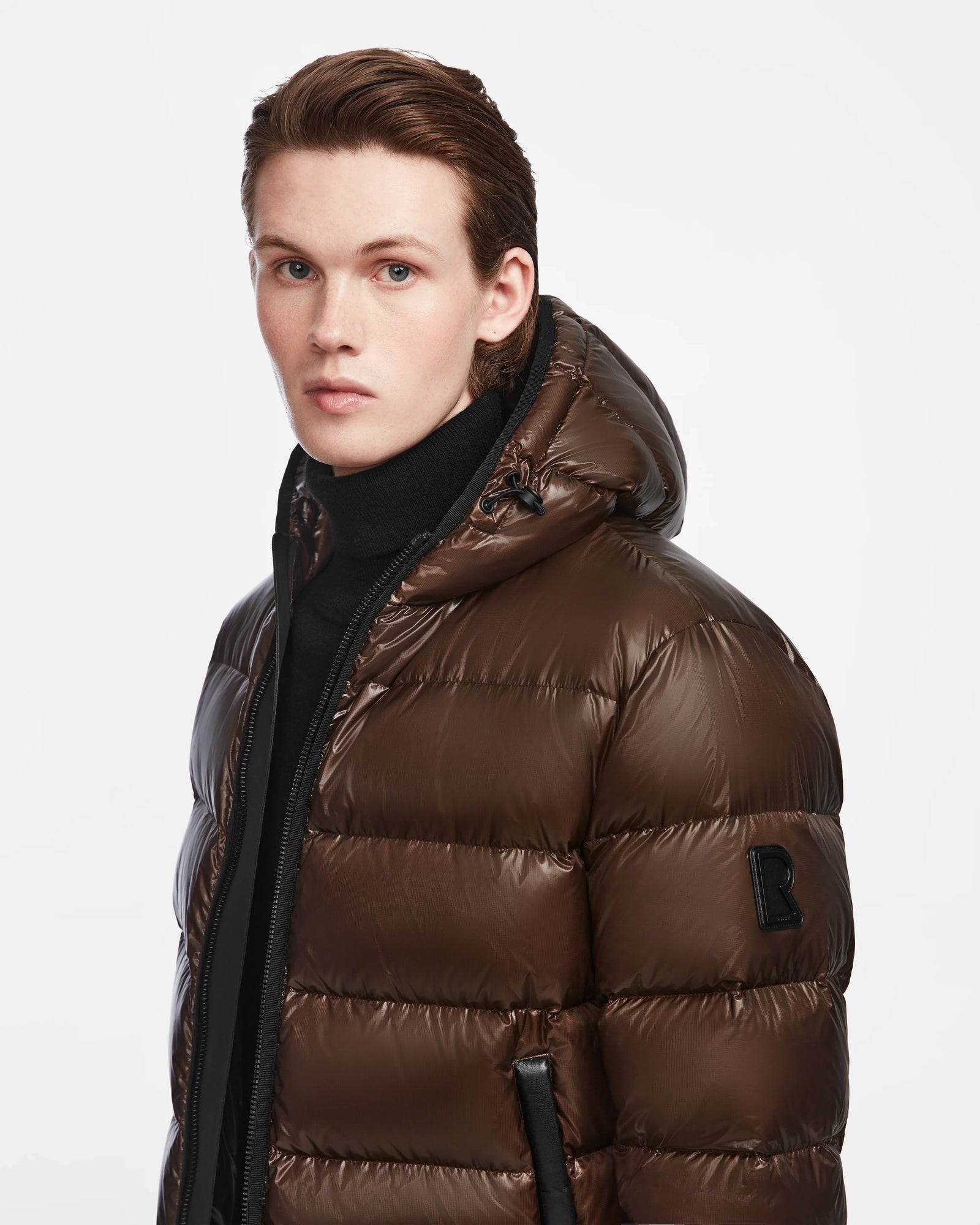 A young person with fair skin and brown hair wears a Rudsak DEMIAN S - M123508 Espresso mens down puffer jacket with hood, styled over a black turtleneck, standing against a white background. The jacket is water and wind resistant for warmth and style.