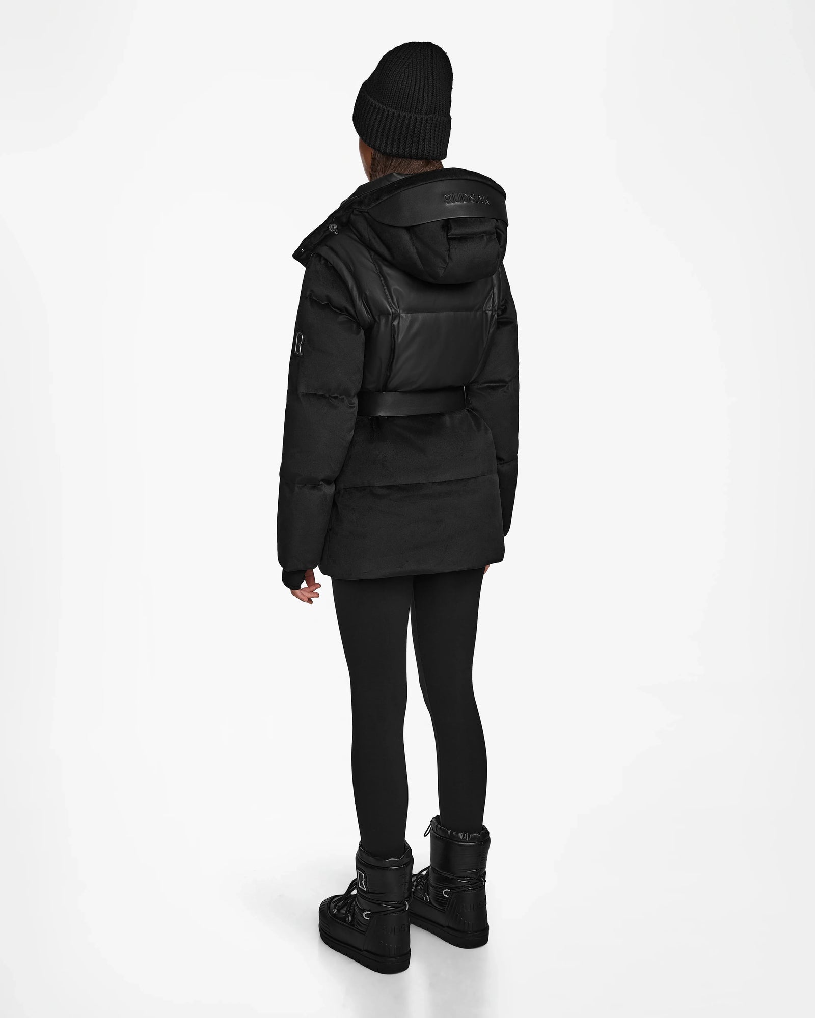 A person stands facing away, wearing the Rudsak DONIA - L124563 Black womens down puffer with leggings, platform boots, and a black beanie against a plain white background.