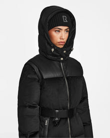 A person wearing the Rudsak DONIA - L124563 Black convertible winter coat with a belted waist and a black knit beanie, looking serious against a plain white background.