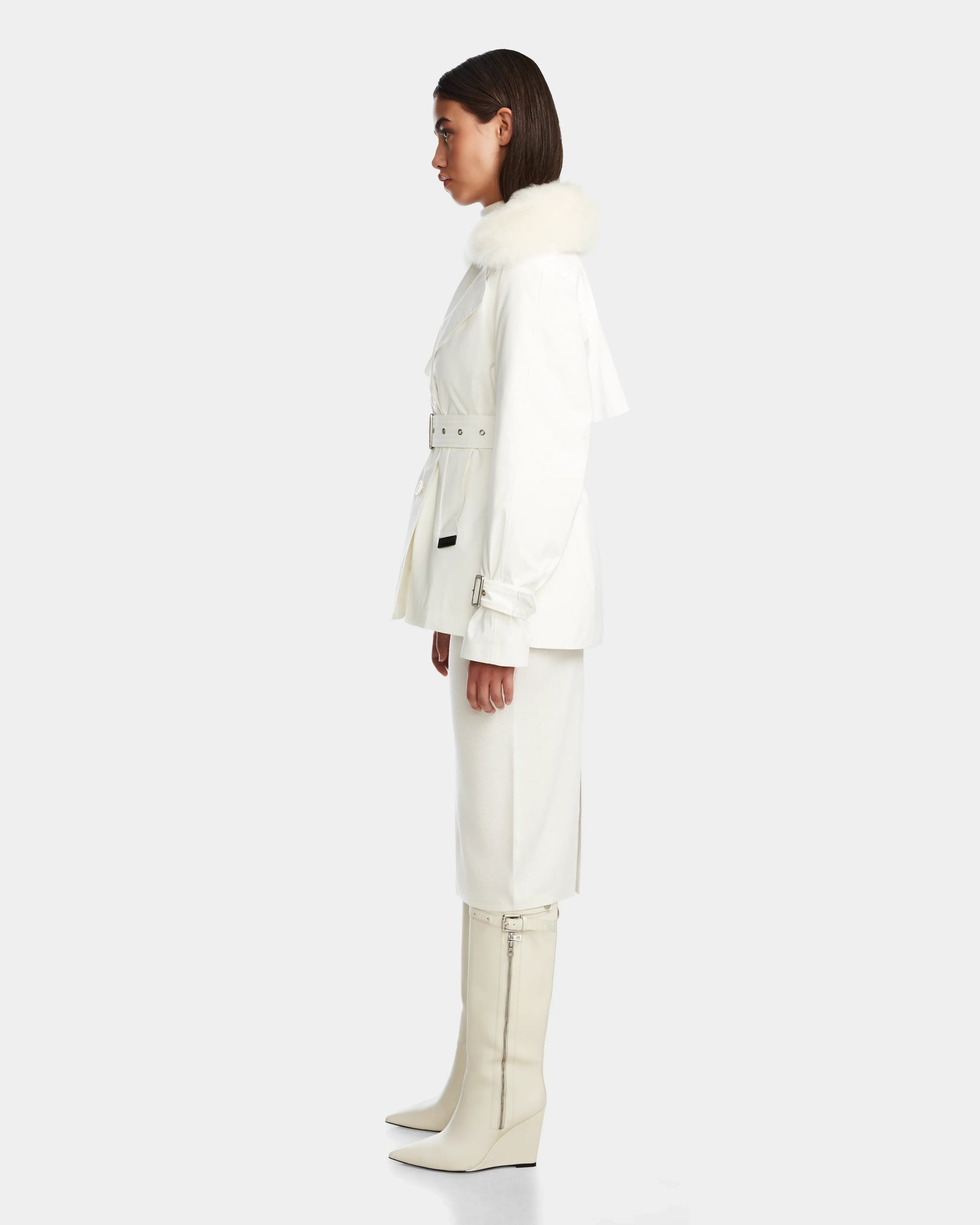 A woman stands in profile against a light gray background, wearing the Rudsak ELENOR - 8124707 Iceland: a women’s all-white trench coat with a shearling collar, styled with a matching skirt and knee-high pointed-toe boots.