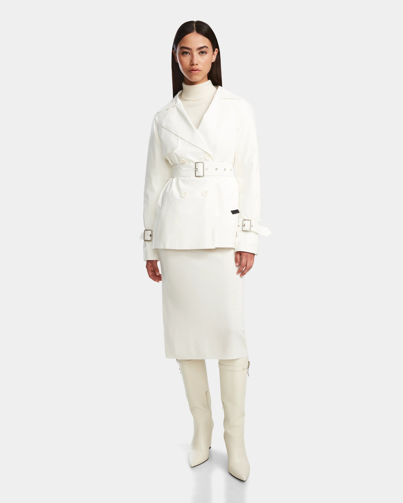 A woman stands against a plain light background wearing the Rudsak ELENOR - 8124707 Iceland trench coat with a shearling collar, a cream turtleneck, matching midi skirt, and white knee-high heeled boots.