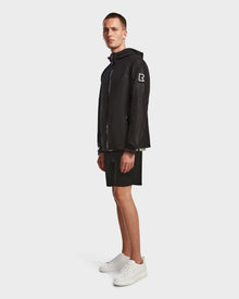 A man stands sideways against a light background wearing the Rudsak ELIJAH - M124000 Black jacket with a white R logo on the sleeve, paired with black shorts and white sneakers.