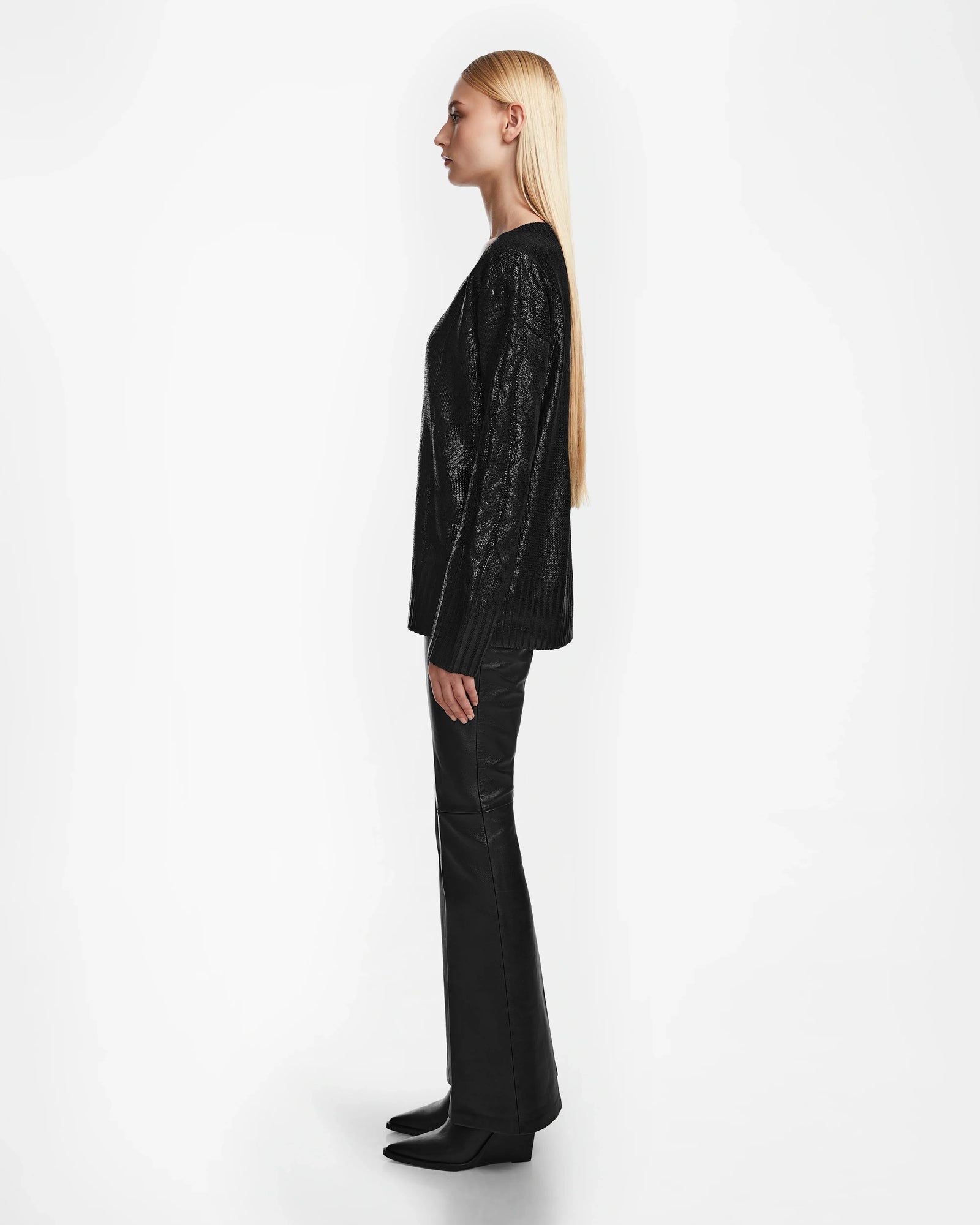 A woman with long straight blonde hair stands in profile, wearing the Rudsak ELLE - 8524597 Black V-neck sweater, black pants, and boots, set against a plain white background.