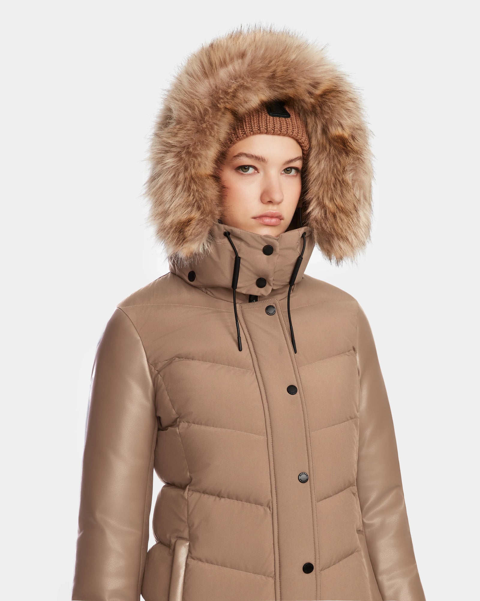 A woman wears the Rudsak ELSA - L123535 Sand quilted down puffer with a faux fur-lined hood and a matching knit beanie, standing against a plain white background.