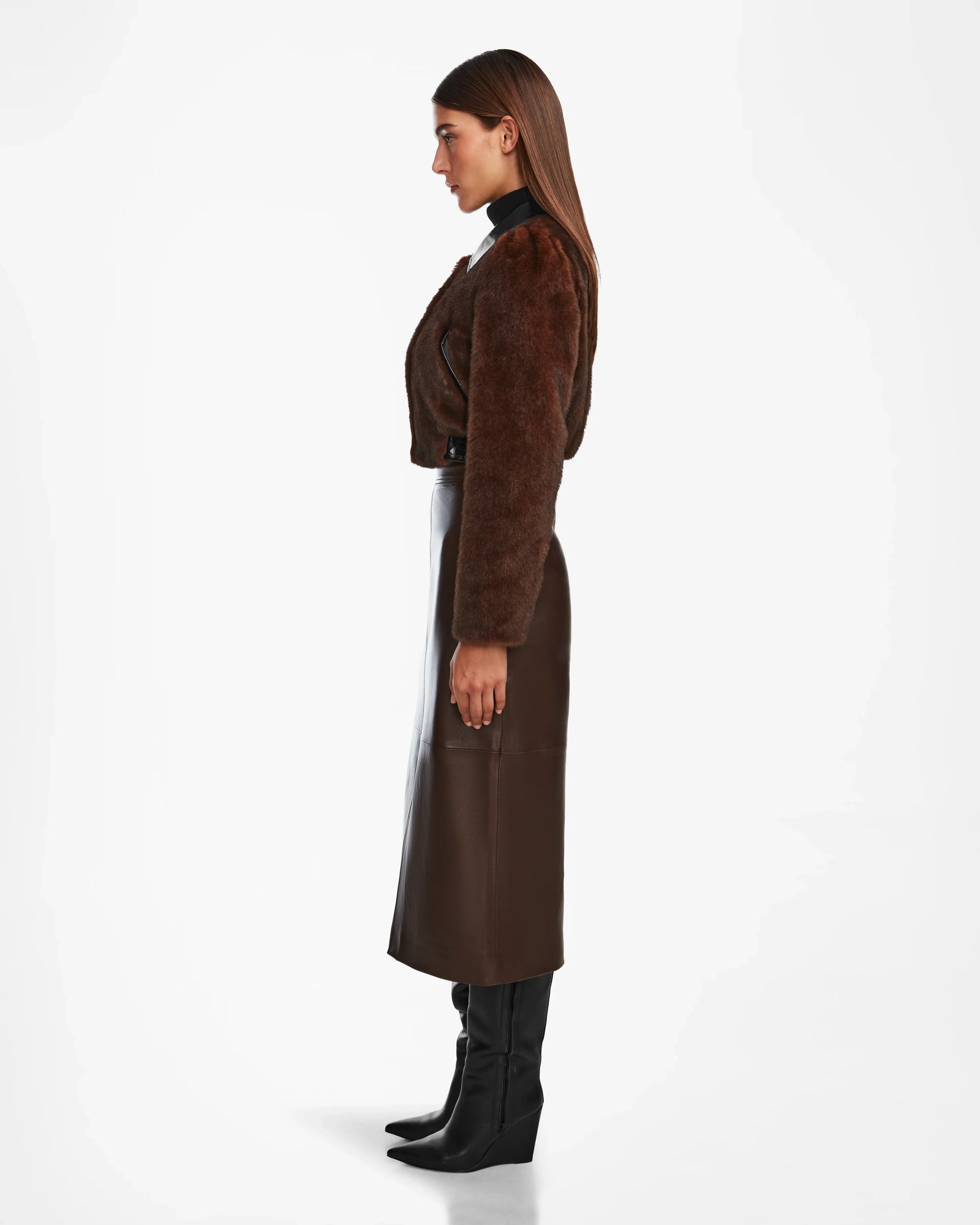 A woman stands in profile against a white background, wearing Rudsaks EVELYN - 8124791 Espresso faux fur jacket, paired with a long dark brown leather skirt and black knee-high heeled boots.