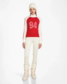 A woman stands against a plain white background wearing the Rudsak FANNY - 8524506 Flame Red après-ski sweater with “84” on the front, paired with a white beanie, white fitted pants, and snow boots.