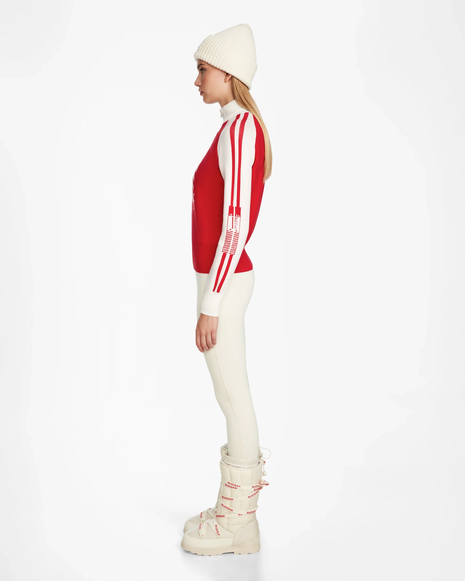 Woman in profile wearing the Rudsak FANNY - 8524506 Flame Red après-ski sweater, a white beanie, cream leggings, and white boots, posed against a plain white background.