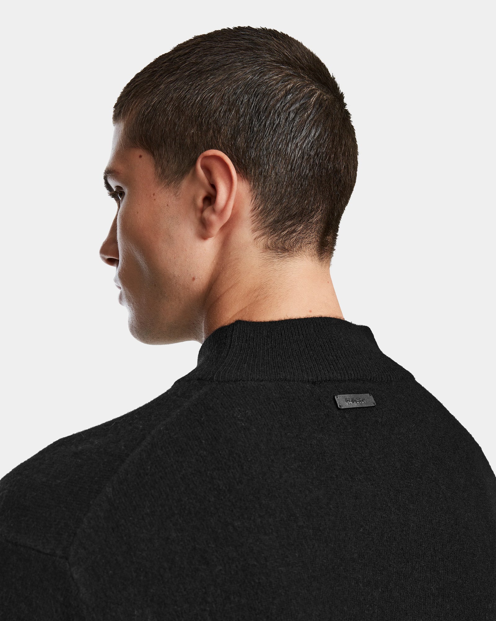 A person with short brown hair is shown from the back and side, modeling the Rudsak FERRAN - 6524511 Black sweater against a plain white background. A small rectangular black tag appears on the upper back of the sweater.