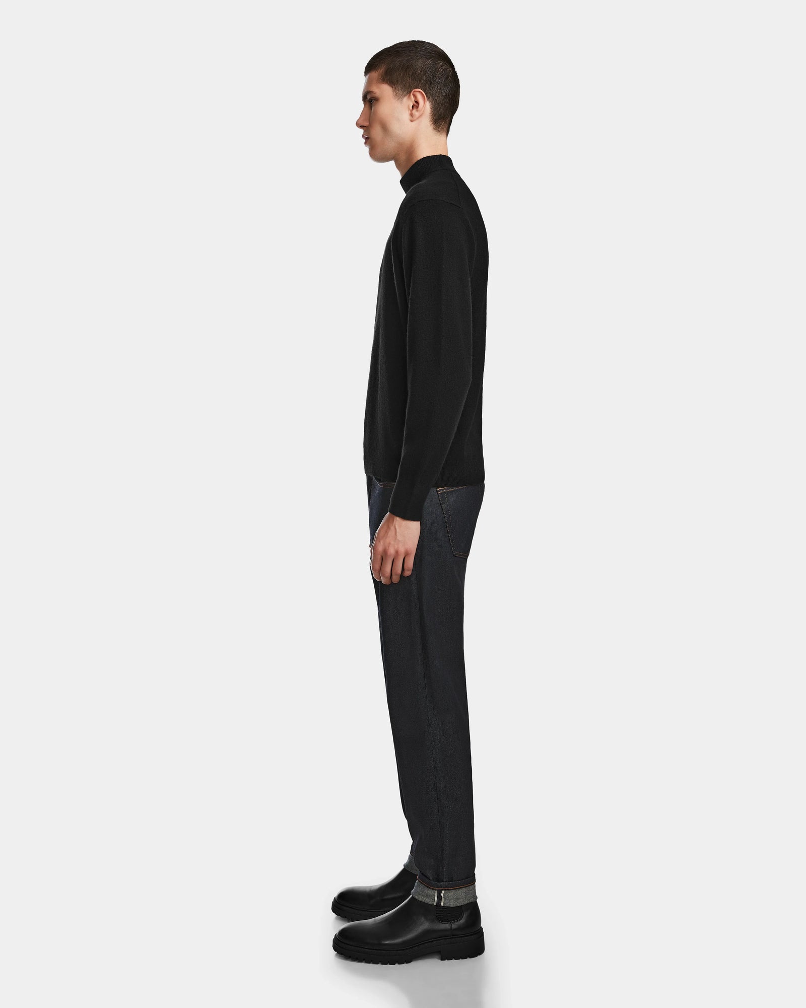 A man stands in profile against a light background, wearing the Rudsak FERRAN - 6524511 Black mens turtleneck sweater, crafted from a luxurious wool and cashmere blend, paired with dark jeans and black boots.