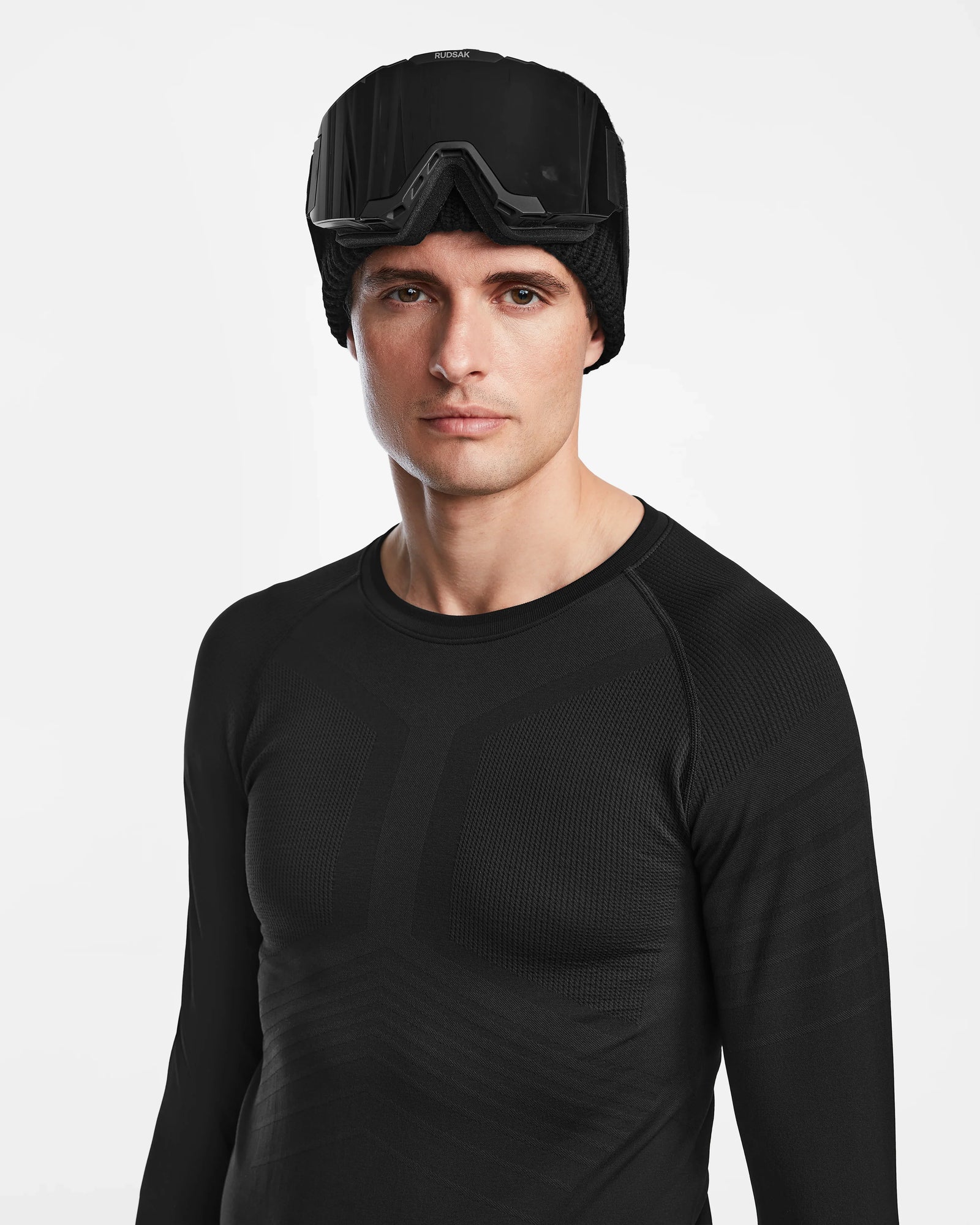A man wearing the Rudsak FRANC - 6524541 Black stretch base layer top, a black beanie, and black ski goggles on his forehead stands against a plain white background.