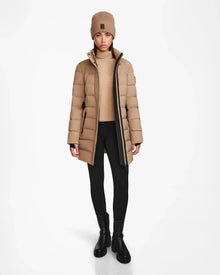 A person stands facing forward wearing the Rudsak FRIDA M - L124524 Sand womens beige mid-length down jacket with a removable hood, styled with a knit beanie, tan sweater, black leggings, and boots against a white background.