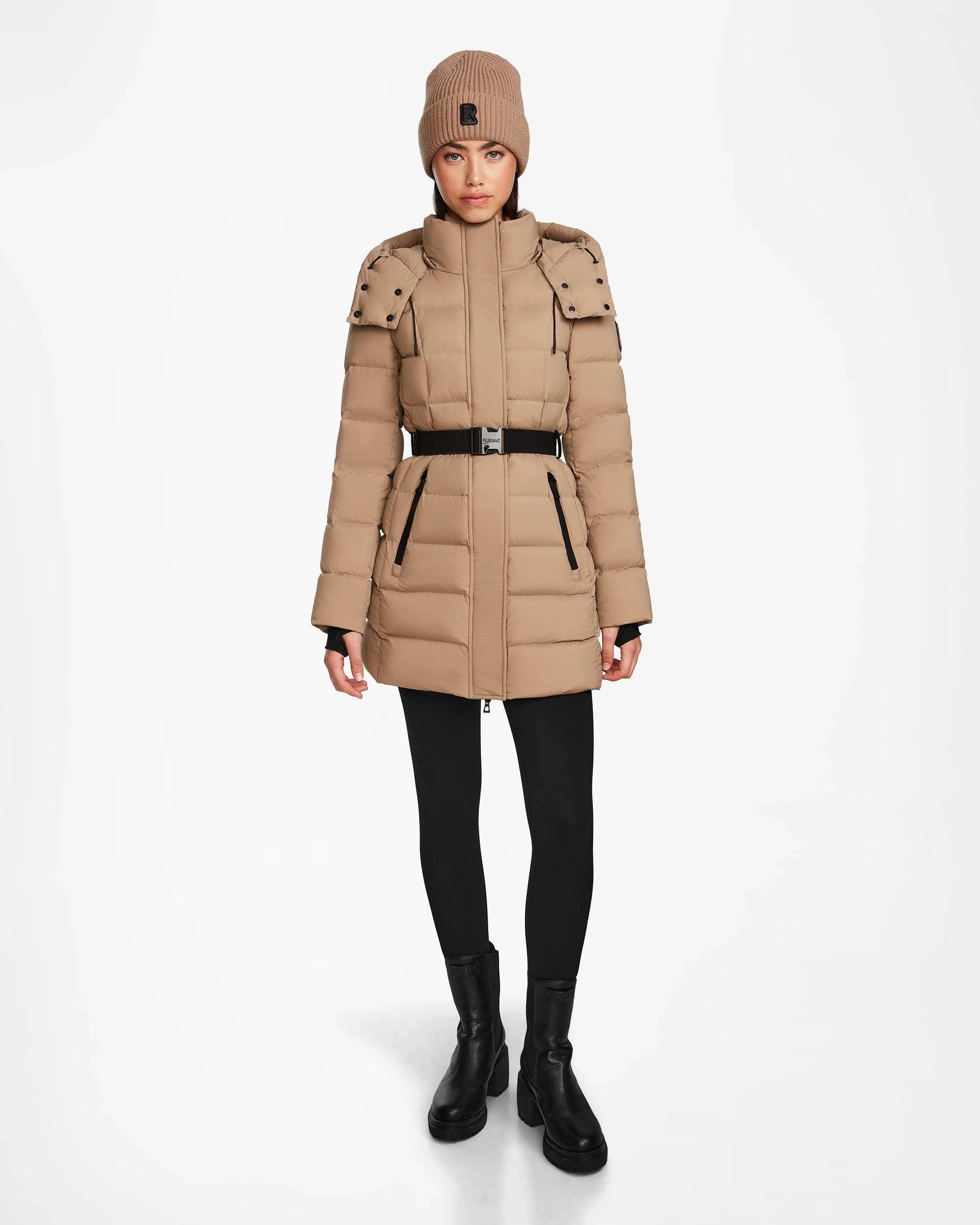 A woman stands facing forward against a plain white background, wearing the Rudsak FRIDA M - L124524 Sand mid-length down jacket along with a matching tan knit beanie, black leggings, and black ankle boots.