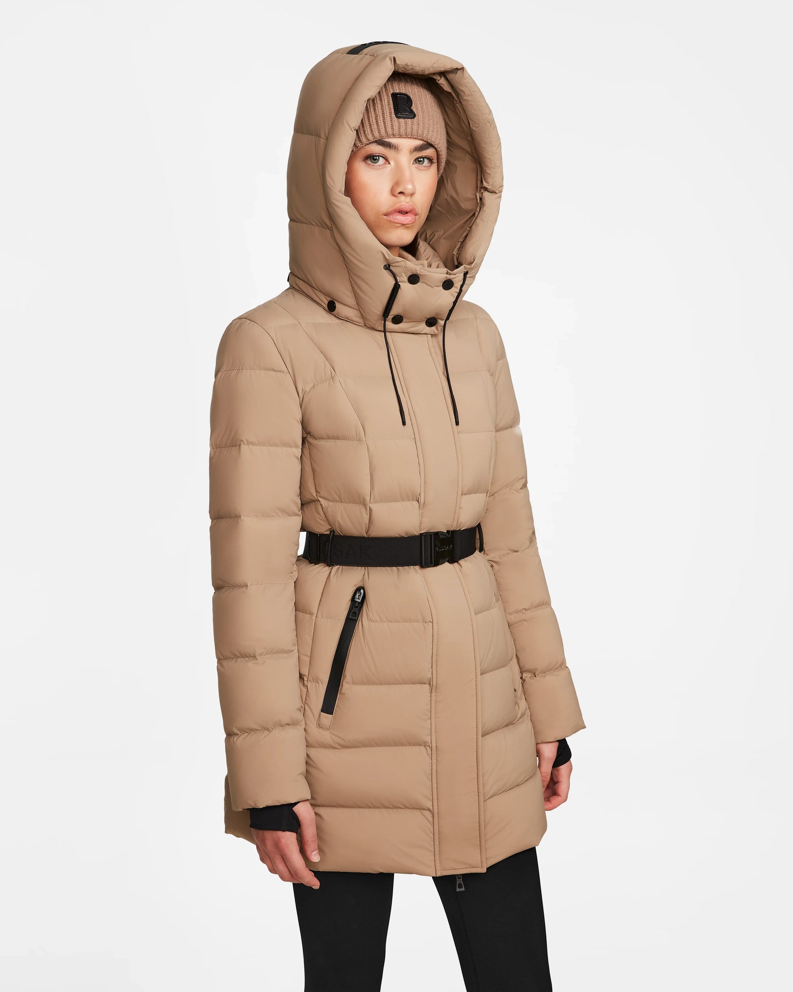 A woman wearing the Rudsak FRIDA M - L124524 Sand, a tan belted mid-length down jacket with removable hood and matching knit beanie, stands facing forward against a plain white background.