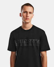 A young man with short hair models the Rudsak GIANNI - 6524535 Black men’s t-shirt, featuring a bold BUDSAK logo in large dark letters across the chest, standing before a plain light background.