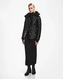 A woman with straight blonde hair and a neutral expression stands against a white background wearing the Rudsak HANNA X - 8123558 Black luxury down jacket, a long black dress, and black boots.