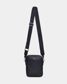 The Rudsak HUBERT - M324500 Black is a black leather crossbody bag with a zippered front pocket and an adjustable fabric strap embossed with RUDSAK, shown against a plain white background.