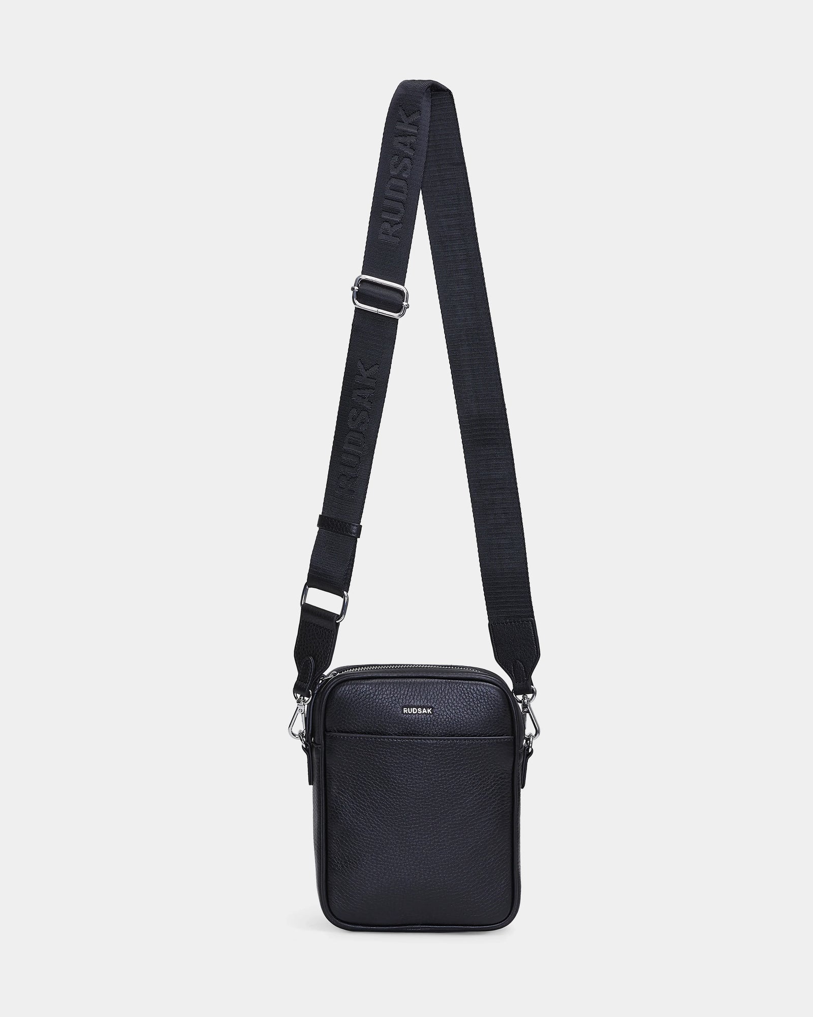 The Rudsak HUBERT - M324500 Black is a black leather crossbody bag with a zippered front pocket and an adjustable fabric strap embossed with RUDSAK, shown against a plain white background.