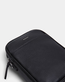 A close-up of the Rudsak HUBERT - M324500 Black leather pouch, featuring a front pocket, visible zipper, and subtle grain texture against a plain white background.