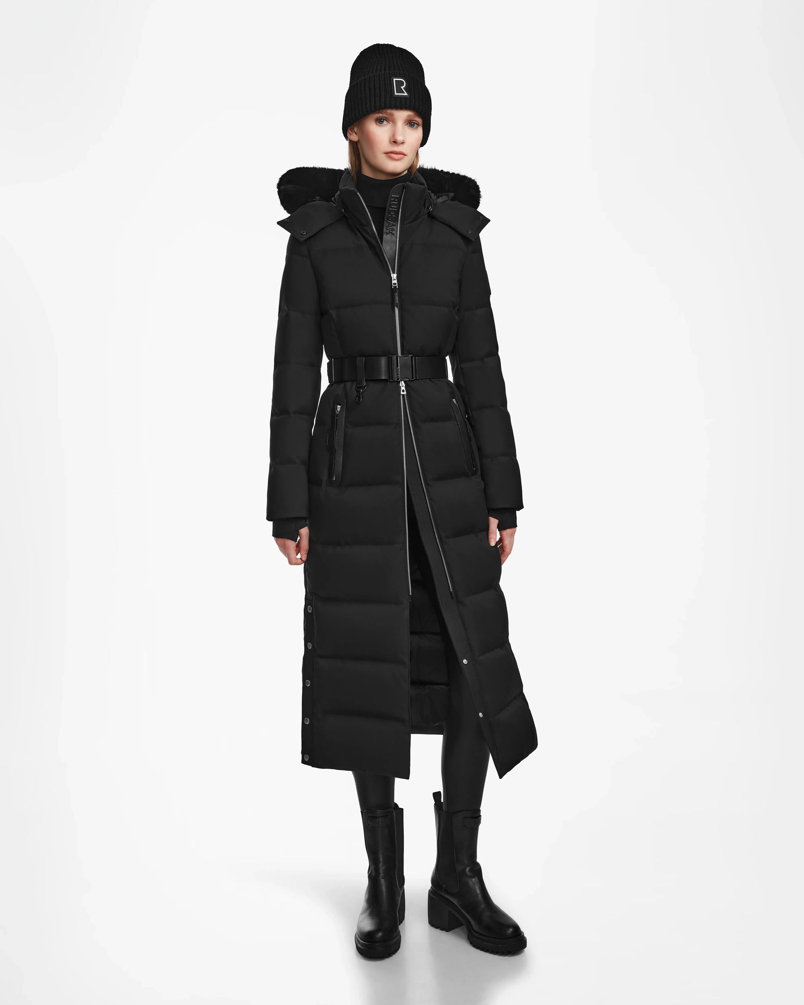 A woman faces forward against a plain white background, modeling the Rudsak IZABELLA - L124541 Black quilted maxi coat with a belt, paired with black boots, leggings, a beanie, and a dark turtleneck—highlighting this winter essential.
