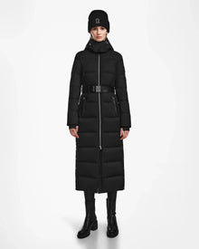 A person stands facing forward wearing the Rudsak IZABELLA - L124541 Black women's long down coat, paired with a black beanie, black pants, and ankle boots against a plain white background.