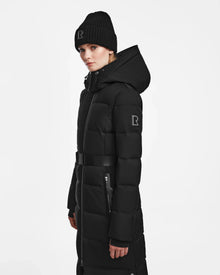 A person stands against a plain white background wearing the Rudsak IZABELLA - L124541 Black women’s down maxi coat—a long, belted black quilted coat with hood—plus a knit beanie, both featuring the signature rectangular R logo. A true winter essential.