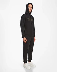 A person stands against a white background wearing the Rudsak JARED - 6524533 Black mens hoodie with the hood up, paired with matching black pants and white sneakers, facing slightly to the side.