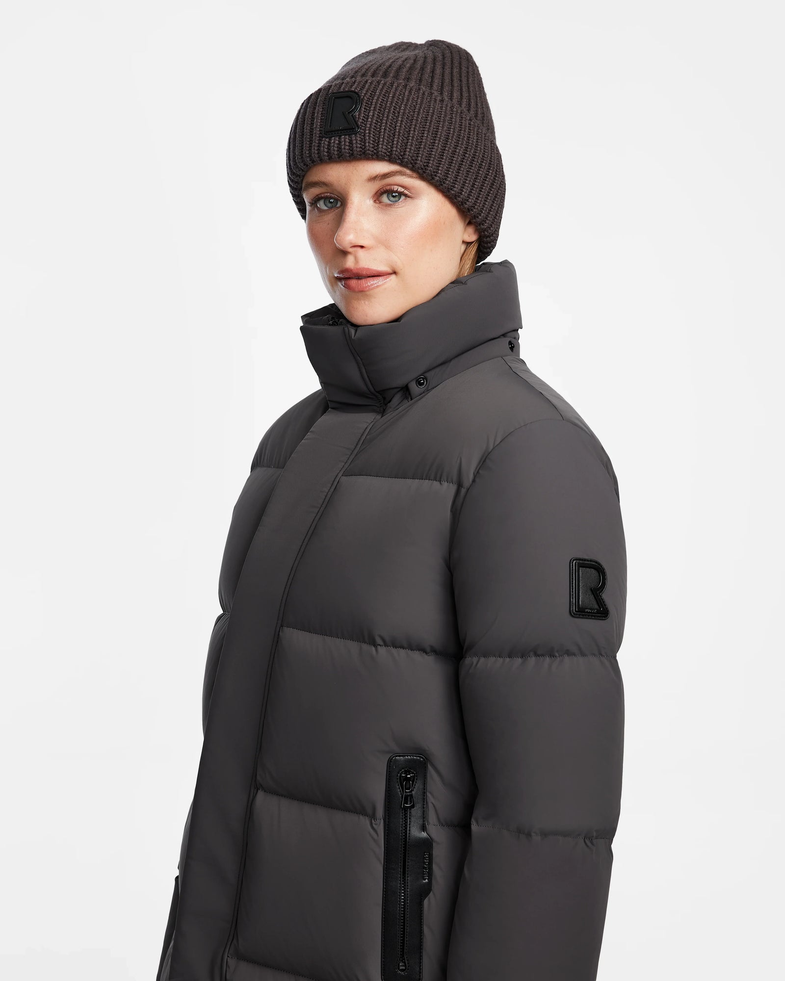 A woman wears the Rudsak JOELLE - 8124729 Grey quilted down coat with a matching knit beanie, both featuring a large R logo and filled with RDS-certified down, standing against a white background and smiling at the camera.