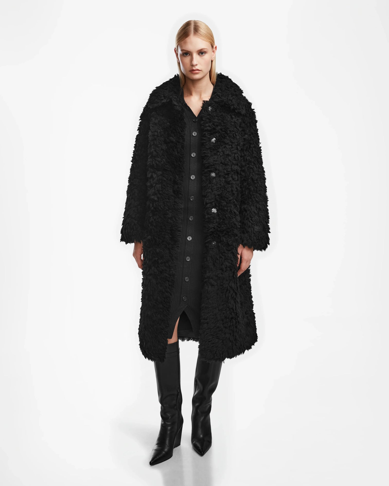 A woman with straight blonde hair and a neutral expression wears the Rudsak KAMILAH - 8124827 Black wool teddy coat over a buttoned black dress with a vegan leather belt and knee-high black boots against a white background.