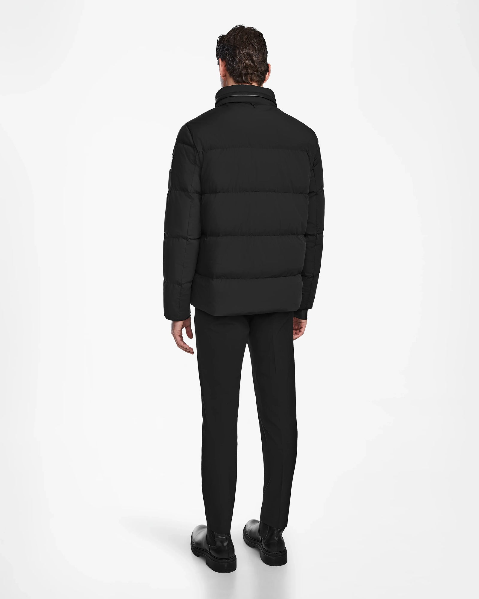 A man faces away, wearing the Rudsak KARLS - M124505 Black men’s puffer jacket with RDS-certified down, paired with black pants and shoes against a plain white background.