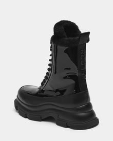 The Rudsak LANA - 8223506 BLACK is a black shiny patent leather mid-calf lace-up boot with a chunky sole, black shearling lining, and a branded pull tab at the back, shown against a plain white background.