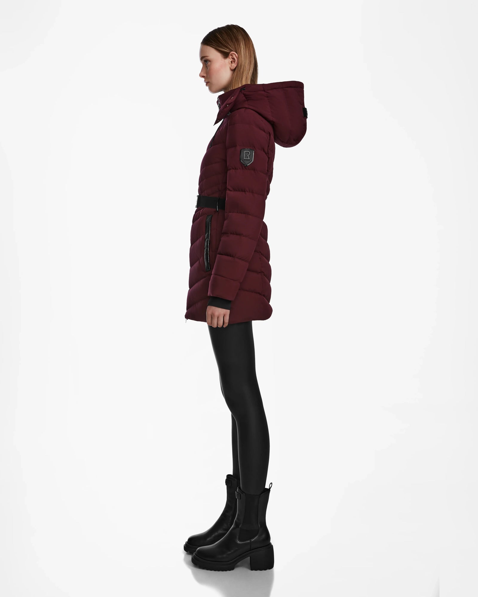 A woman in profile wears the Rudsak MALAYA M - L123554 Wine down puffer jacket with a hood, black belt, black leggings, and black ankle boots against a white background.