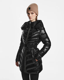 A woman wearing the Rudsak MALAYA S - L123510 Black shiny down puffer jacket with a belted waist and a beige knit beanie stands against a white background, looking slightly to the side with a neutral expression.