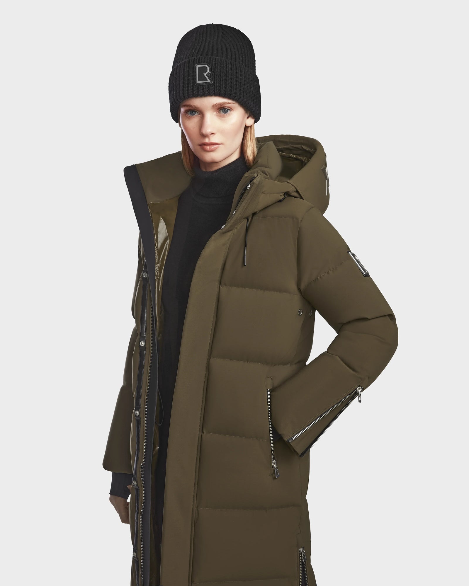 A woman faces forward against a white background, wearing the Rudsak MAO M - 8123598 Army womens long olive-green down puffer coat with a hood in weatherproof recycled Stormshell material and a black knit beanie.