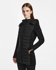 A woman with straight brown hair wears the Rudsak MARRY M - L123548 Black puffer jacket, featuring quilted panels, zippered pockets, and a removable hood. She stands against a plain white background with arms at her sides and a neutral expression.