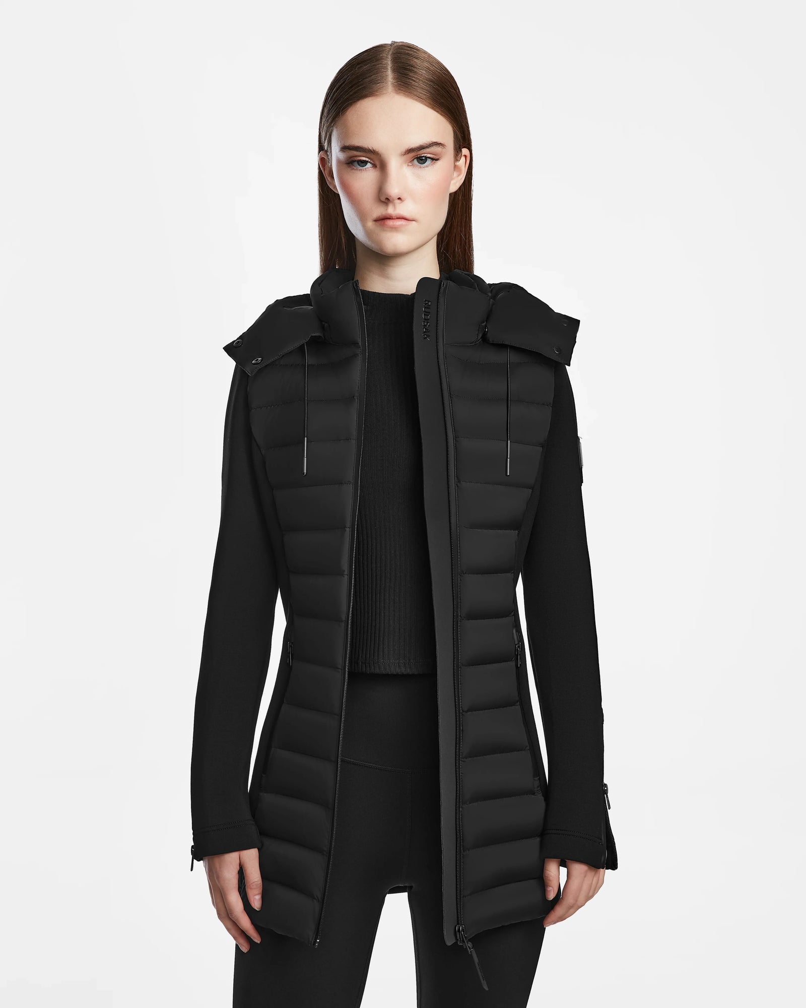 A woman with straight brown hair wears the Rudsak MARRY M - L123548 Black women’s puffer jacket, featuring a removable hood, over a black sweater and leggings against a plain white background.