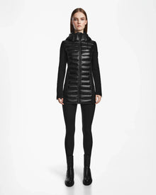 A woman stands facing forward against a plain white background, wearing the Rudsak MARRY - L121003 BLACK recycled down women’s puffer jacket with a hood, paired with black leggings and black ankle boots.