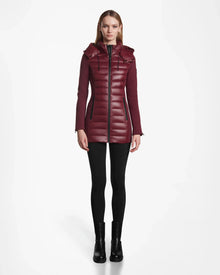 A woman faces forward, wearing the Rudsak MARRY - L121003 Wine long puffer with hood, black leggings, and black ankle boots against a plain white background.