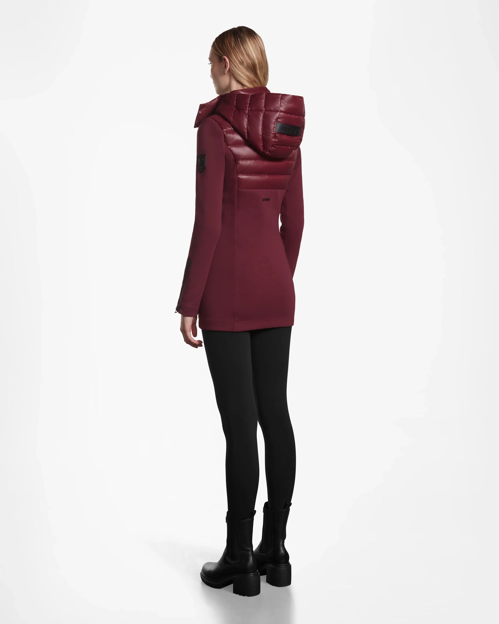 A person with blond hair faces away, wearing the Rudsak MARRY - L121003 Wine women’s puffer with padded hood, paired with black leggings and boots against a plain white background.