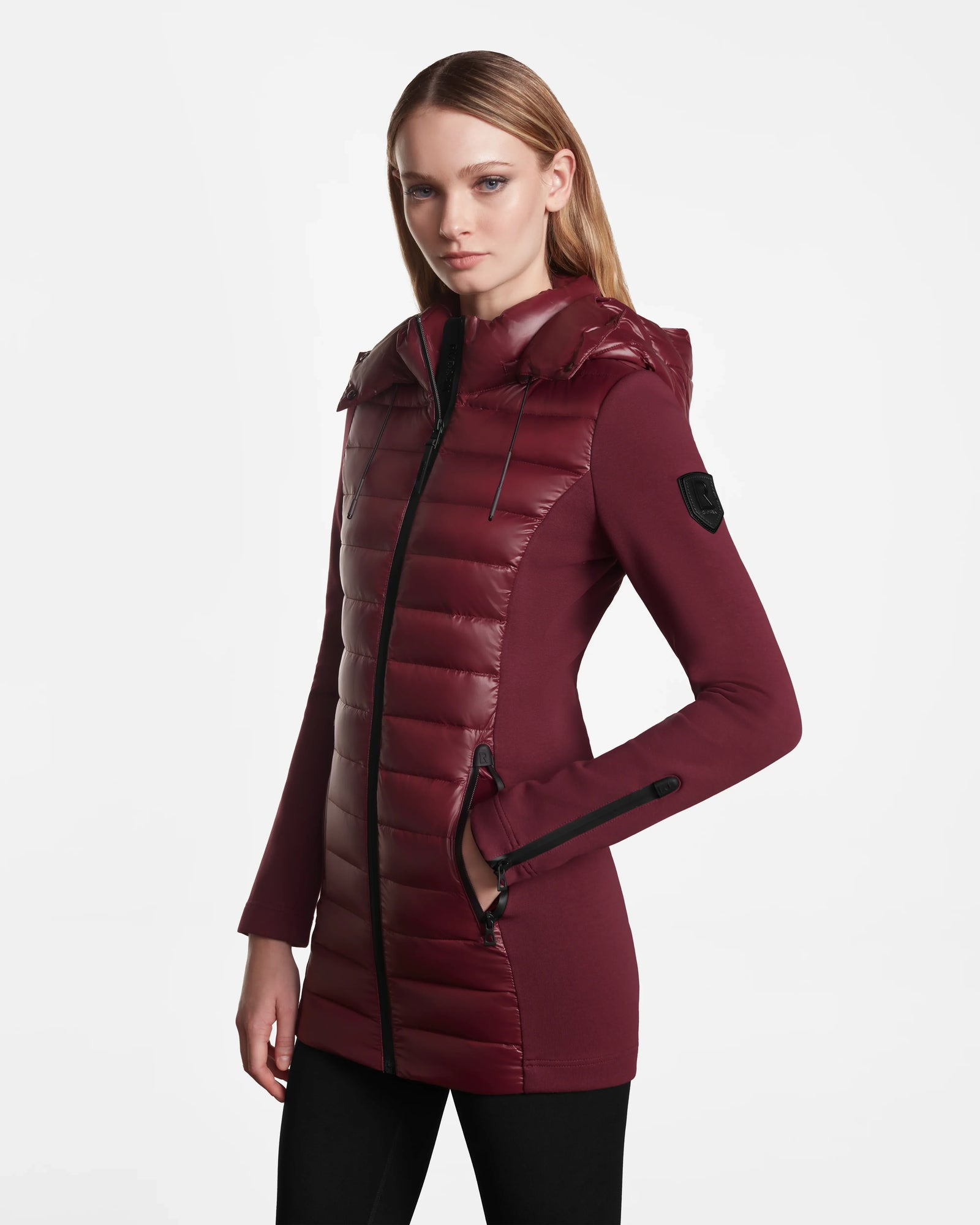 A woman in a Rudsak MARRY - L121003 Wine activewear-inspired maroon hooded puffer jacket with fitted sleeves and zip pockets, standing against a plain white background.