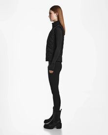 A woman in profile stands against a white background, wearing the Rudsak MATILDA - L124504 Black vest over a black turtleneck, leggings, and ankle boots. Her straight brown hair is down—a chic look for transitional weather.