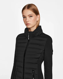 A young woman with straight, light brown hair wears the RUDSAK MERINA - 8122546 BLACK quilted down jacket, featuring a zip-up front and high collar. She stands against a plain white background, gazing slightly to the side.
