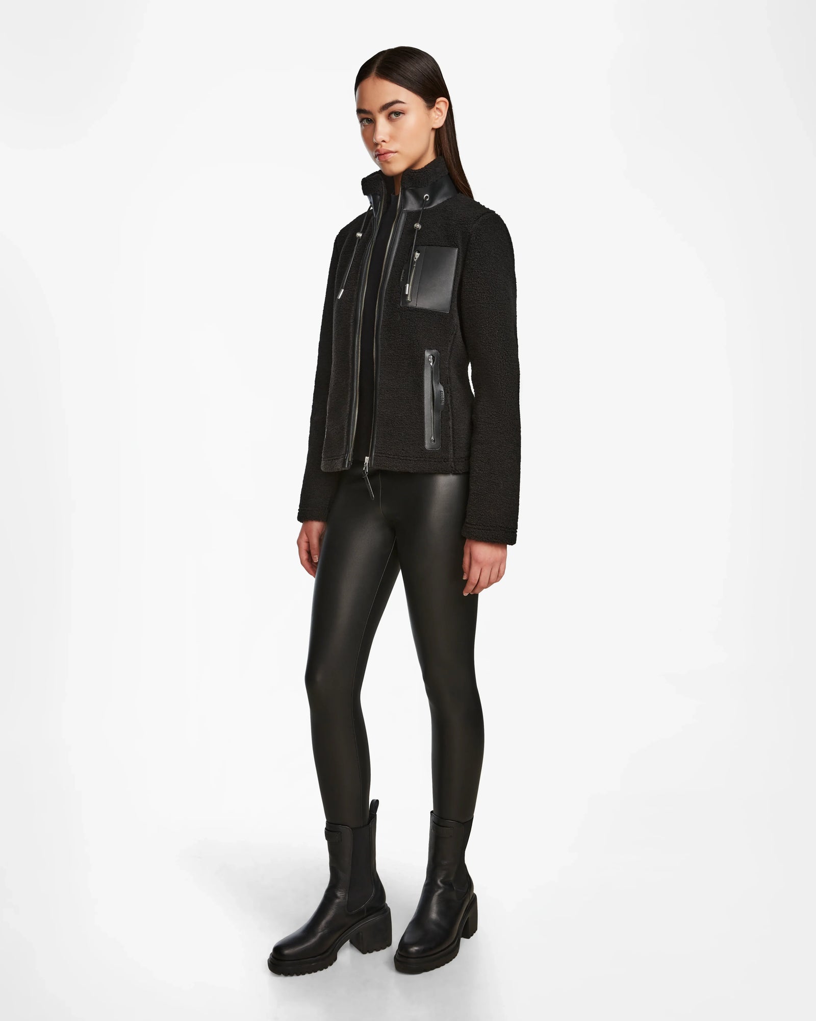 A woman faces slightly to the side wearing Rudsaks MILANA - 8524604 Black zip-up jacket with vegan leather accents, leather-look leggings, and chunky black boots against a plain white background.