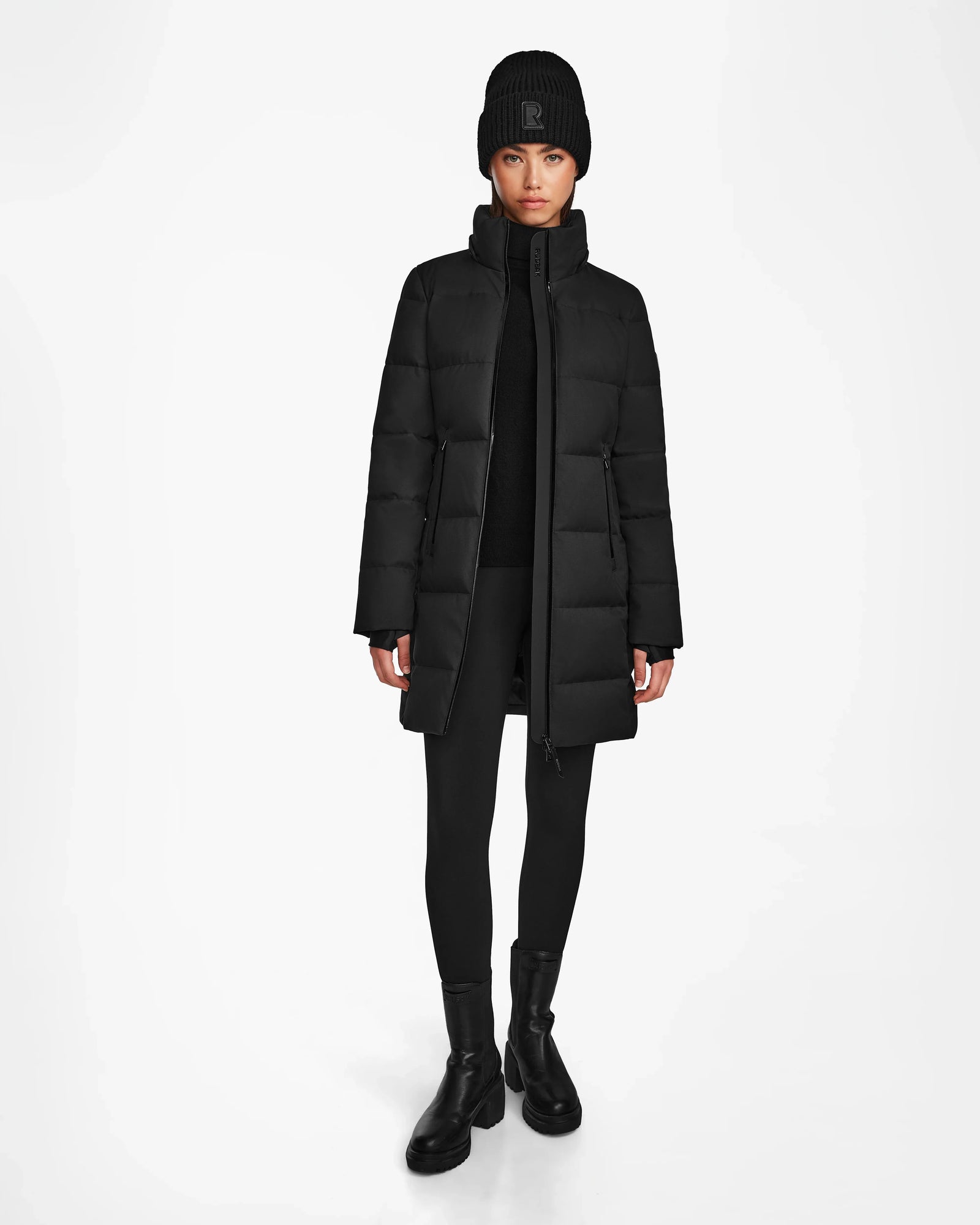 A person stands facing forward, wearing the Rudsak MIRIAM - L122523 BLACK women’s down puffer coat, paired with a black knit hat, leggings, and boots against a plain white background.