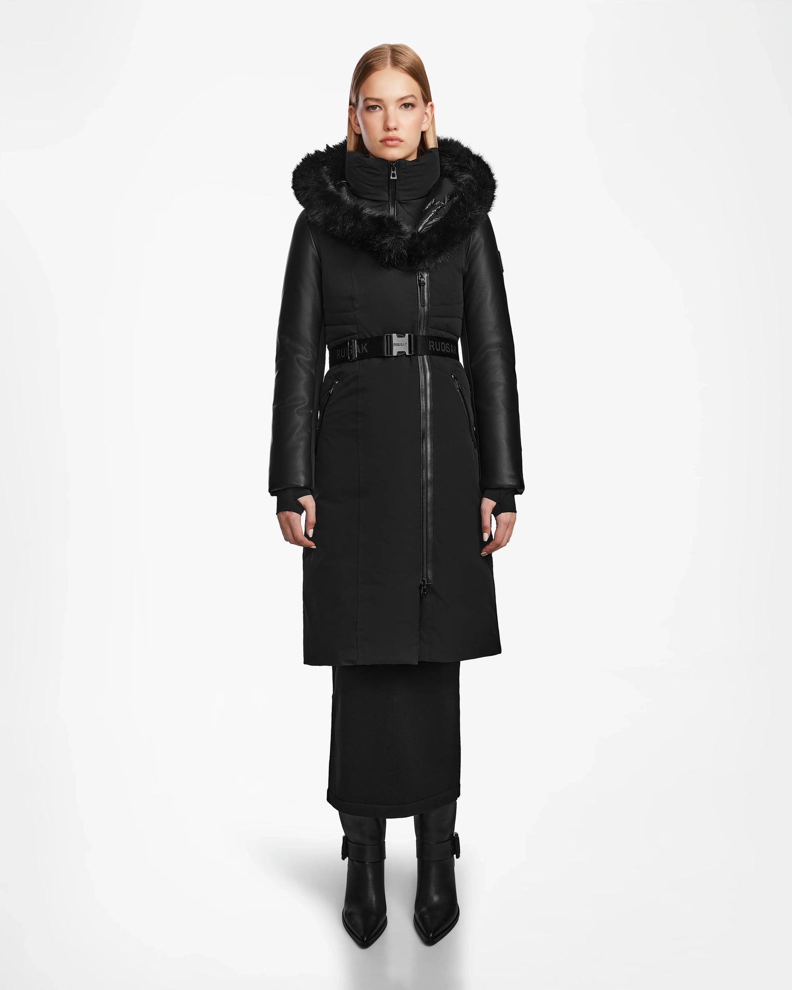 A woman in a Rudsak MISALA - L124512 Black water-resistant down coat with a fur-lined hood and belt stands against a white background, her straight blonde hair and black heeled boots complementing the stylish winter look.