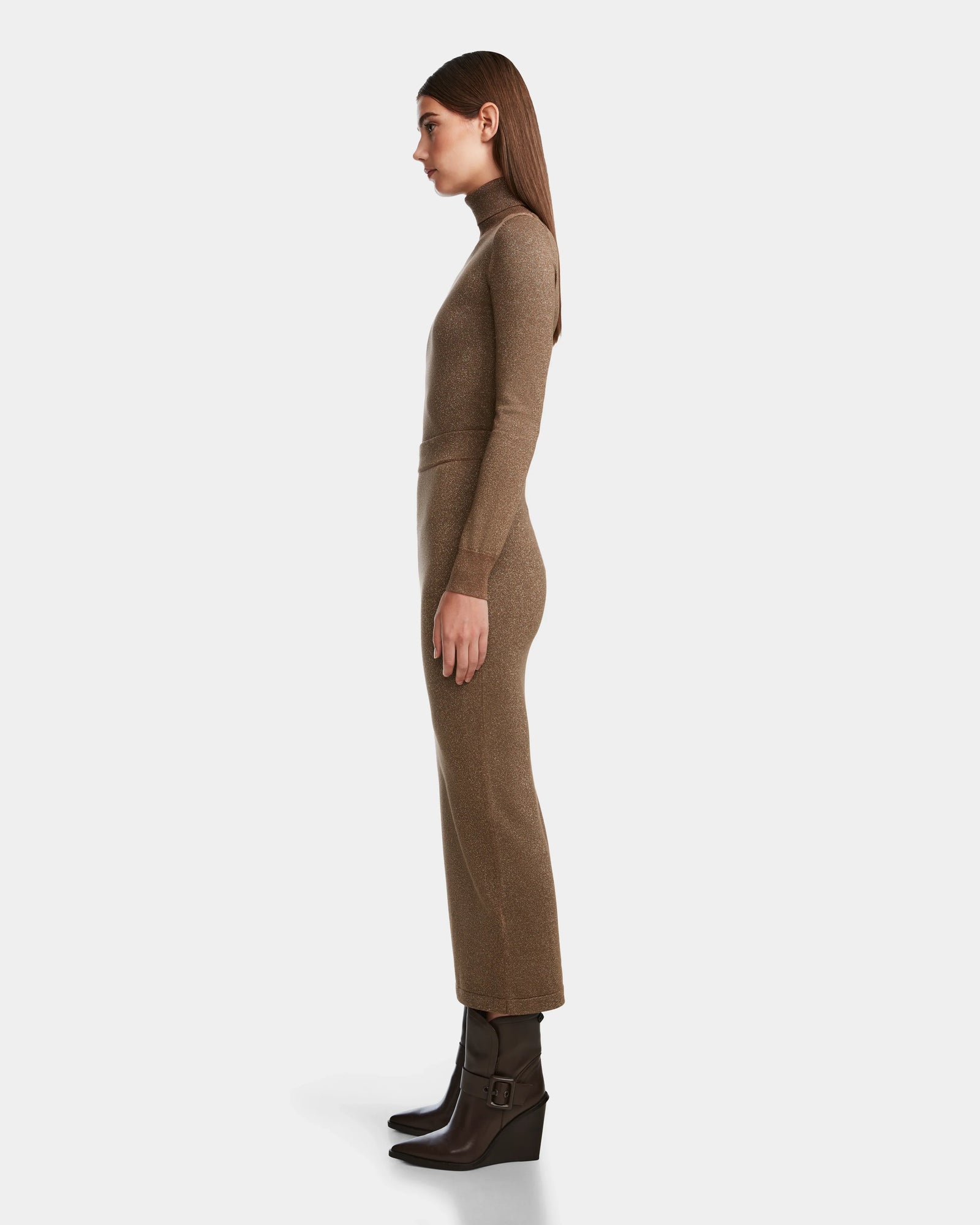 A woman in profile stands against a white background, wearing the MONICA - 8524516 Antique Bronze long-sleeve knit dress by Rudsak, featuring a body-hugging fit, high neckline, ankle-length hem, and paired with brown buckle-strap ankle boots.