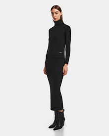 A woman with straight brown hair and a neutral expression poses against a plain background wearing Rudsaks MONICA - 8524516 Black fitted turtleneck, a matching knit skirt, and black heeled ankle boots.