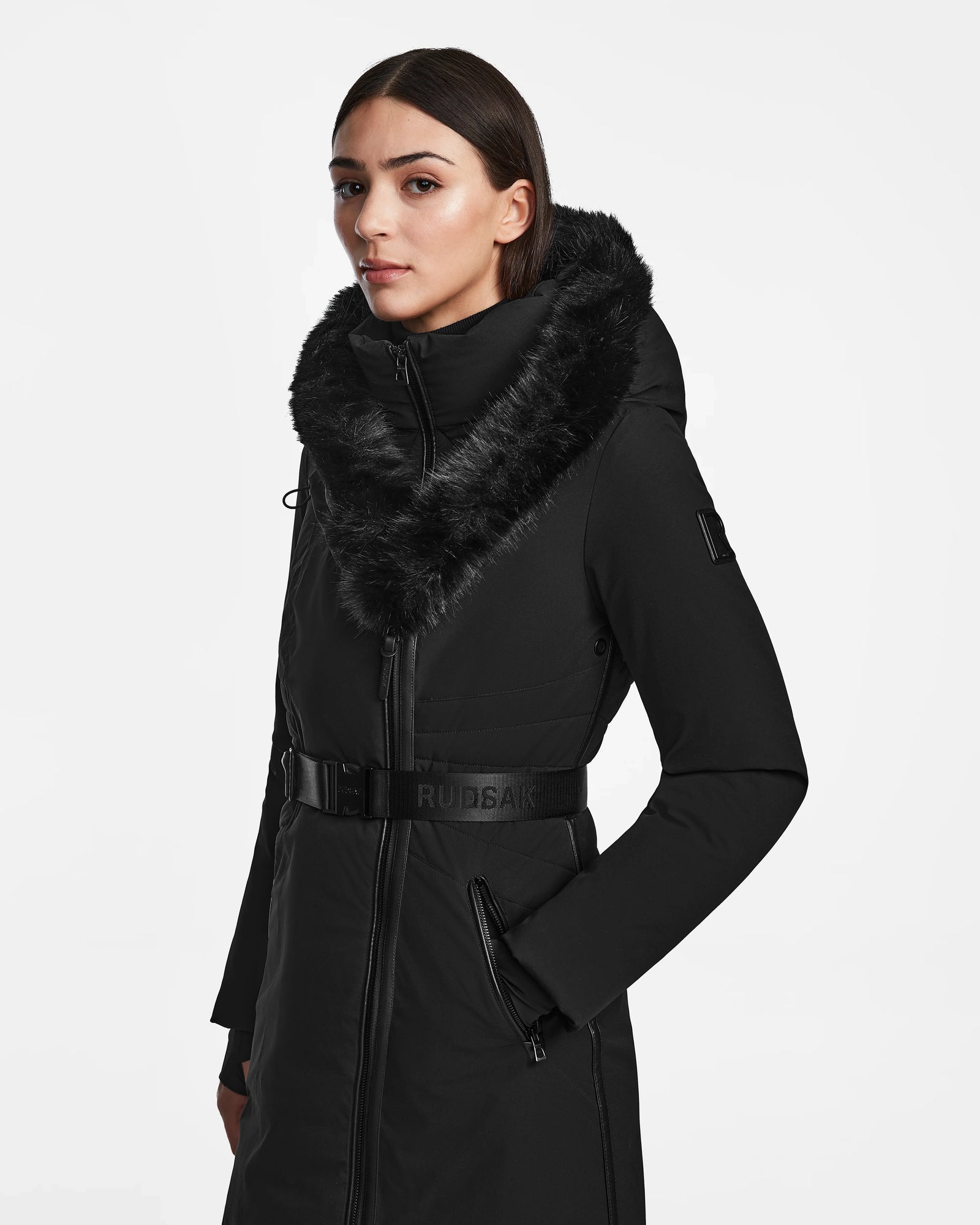 A woman wearing the Rudsak MUSA - L124534 Black maxi down puffer with a belted waist, fur-lined hood, and front zipper—made from recycled stretch shell and RDS-certified down—stands against a plain white background.
