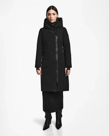 A woman stands facing forward against a white background, wearing the Rudsak MUSA - L124534 Black long down puffer with a high collar and asymmetrical zipper, paired with a black skirt and heeled boots.